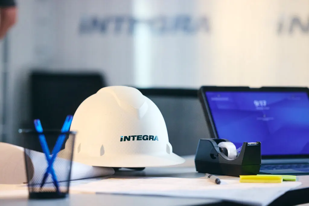 An Integra hard hat resting on a desk beside drawings and a laptop